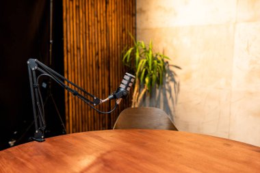 Bir podcast stüdyosunda ahşap bir masanın yanına yerleştirilmiş şık bir mikrofon bulunur. Kurulum doğal ışıkla aydınlanıyor ve kayıt oturumları için davetkar bir atmosfer yaratıyor..