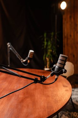 Konforlu bir stüdyo, ahşap bir masanın üzerine yerleştirilmiş sağlam mikrofonlar içeren bir podcast kaydı için hazırlanıyor. Ortam sıcak, sohbet etmek için ideal..