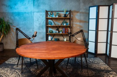 Kollara bağlı iki profesyonel mikrofonun yer aldığı bir podcast seansı için yuvarlak ahşap masa hazırlanıyor..