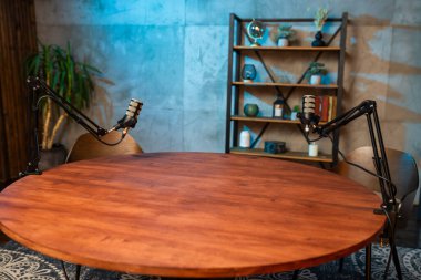 Mobilyalı bir podcast stüdyosu ayarlanabilir mikrofonlarla donatılmış yuvarlak ahşap bir masa sergiliyor. Şık bir kitaplık ve yeşillik yaratıcı atmosferi geliştirir..