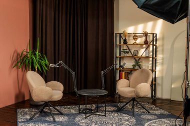 Konforlu podcast stüdyosunda, ayarlanabilir mikrofonlarla eşleştirilmiş, küçük bir masanın karşısına iki davet edici sandalye sergileniyor. Bir saksı bitkisi ve dekoratif raflar sıcaklık ekler..