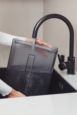 Elleri lavabo musluğunun altında şeffaf bir su filtresi durulandırırken gösteriliyor, böylece su kalitesi ve sağlık güvenliği için kirletici madde ve kirlilikten arınmış oluyor..