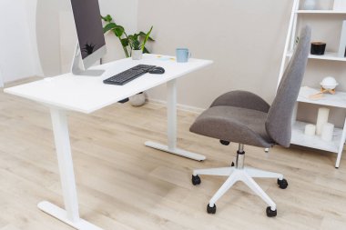 Çağdaş bir ofis gri ergonomik sandalyeyle eşleştirilmiş parlak beyaz bir masaya sahiptir. Bir bilgisayar monitörü, klavye ve dekoratif bitki davet edici bir çalışma alanı oluşturur..