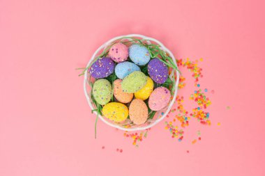 Renkli Paskalya yumurtalarıyla dolu dekoratif bir sepet pembe arka planda duruyor. Sepetin etrafında küçük şekerlerden oluşan bir çeşitlilik var, neşeli bir atmosfer yaratıyor..