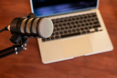 Tahta bir masadaki dizüstü bilgisayarın yanına yerleştirilmiş bir mikrofon aktif bir podcast kaydını gösteriyor. Ortam eğitim ve eğitim için odaklanmış bir ortam öneriyor.