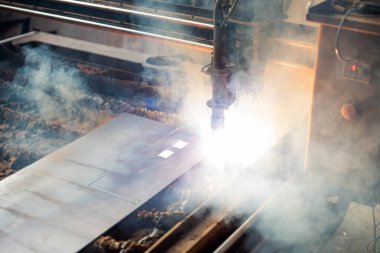 Bir işçi, metal imalat atölyesinde plazma kesme makinesi kullanıyor. Parlak ışık ve duman havayı dolduruyor. Metal tam olarak kesildiği için gelişmiş teknoloji iş başında..