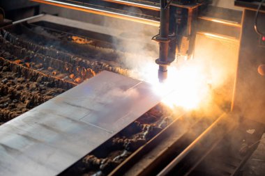 Bir endüstriyel atölyede plazma meşalesi kullanılarak metal bir levha kesiliyor. İşlem, metal üretiminde kesme teknolojisiyle hassasiyeti gösteren kıvılcımlar ve duman üretiyor..