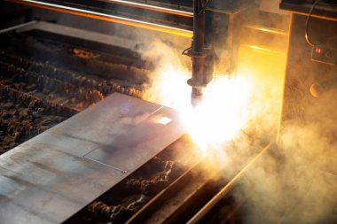 Bir işçi, bir atölyedeki metal bir levhayı kesmek için plazma meşalesi kullanıyor, duman ve kıvılcımlarla çevrili, endüstriyel üretim çalışmalarının hassasiyetini ve yoğunluğunu vurguluyor..