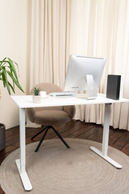 Temiz beyaz bir masa ve rahat bir sandalyenin bulunduğu çağdaş bir çalışma alanı. Masanın üzerinde küçük bir bitki ve dekoratif bir element ile tamamlanmış parlak bir monitör duruyor..