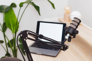 Arka planda dizüstü bilgisayar ve yeşillik bulunan bir masanın üzerine yerleştirilmiş bir mikrofon, çağdaş bir ofiste podcast veya eğitim kayıt oturumuna hazırlık niteliğindedir..