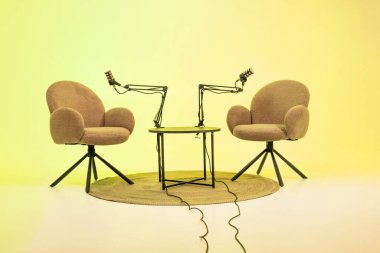 Modern bir podcast stüdyosunda, iki rahat sandalye dairesel bir masanın etrafına yerleştirilmiştir. Her sandalye bir mikrofon ile donatılmıştır, mülakatlar ve eğitimsel konuşmalar için idealdir.