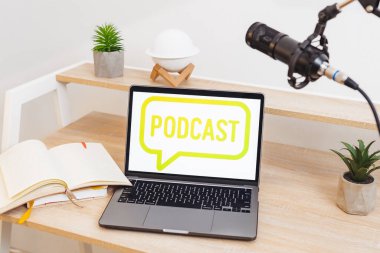 Podcast sözcüğünü gösteren bir dizüstü bilgisayar bir defter ve bitkinin yanında bir masanın üzerinde duruyor. Yakınlarda bir mikrofon var, eğitici bir görüşme ya da konferans için hazır..