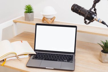 Tahta bir masada, bir mikrofon belirgin bir şekilde bir dizüstü bilgisayar ve açık bir not defterinin yanına yerleştirilir. Bu kurulum, podcast dersleri ve eğitime odaklı röportajlar için tasarlanmıştır..