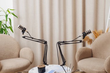 Bir podcast kaydı için ayarlanabilir mikrofonları olan şık bir masada iki pelüş sandalye karşı karşıya. Sakin bir iç mekan atmosferi rahat konuşmaları teşvik eder..