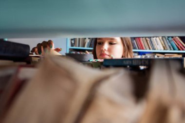 Genç bir öğrenci canlı bir kütüphanedeki bir raftan kitap seçmeye odaklanmış. Onu çevreleyen sayısız kitap öğrenme ve keşif atmosferi yaratıyor..