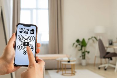 Bir kullanıcı şık bir oturma odasında otururken akıllı ev teknolojisiyle cep telefonunda etkileşim kurar. Modern mekan doğal ışık, yeşil bitkiler ve minimalist dekora sahiptir..