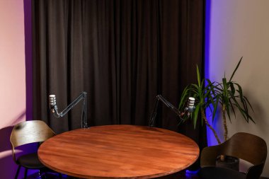 Konforlu podcast stüdyosu yuvarlak bir ahşap masa, iki mikrofon standı ve kulaklıklar içeriyor, canlı yayın ve ilgi çekici tartışmalar için destekleyici bir ortam yaratıyor..