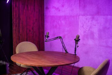 Yuvarlak ahşap bir masa, iki sandalye ve ayarlanabilir kollarda iki mikrofon bulunan modern bir podcast stüdyosu. Ortam aydınlatması kayıt oturumları için sıcak ve davetkar bir atmosfer yaratıyor..