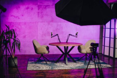 Podcast yayınları için ayrılmış bir alan. İki sandalyeyle çevrili şık bir yuvarlak masa sergileniyor. Renkli ışıklandırma, tartışmalar ve dersler için davetkar bir atmosfer ekler..