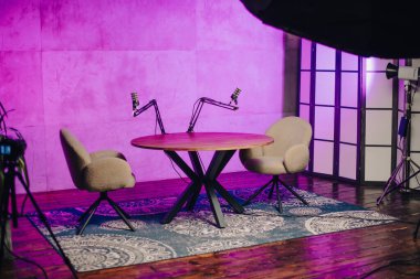 Podcast stüdyosuna iki rahat sandalye ve bir yuvarlak masa hakim. Ayar canlı ışıklarla aydınlatılıyor ve yayın ve tartışmalar için davetkar bir ortam yaratıyor..