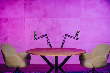 İyi ışıklandırılmış bir stüdyoda, iki rahat sandalye ahşap bir masaya bakıyor ve mikrofonlar canlı yayın için yerleştirilmiş. Renkli arka plan davetkar atmosferi arttırır.