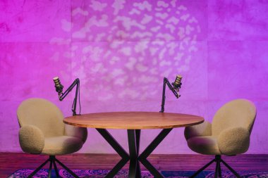 Konforlu bir podcast kayıt alanı yuvarlak bir ahşap masada konumlanmış iki modern sandalye ve mikrofona sahiptir. Ortam, duvardaki renkli ortam aydınlatmasıyla daha da güzelleşiyor..