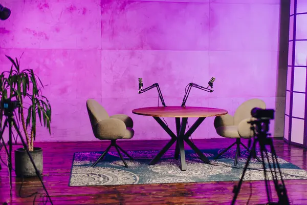 İyi tasarlanmış bir podcast stüdyosunda, yuvarlak ahşap bir masa iki rahat sandalye arasında oturur, mikrofonlar ve kulaklıklarla donatılmış, atmosferi geliştiren renkli ışık altında..