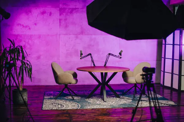 Podcast yayınları için ayrılmış bir alan. İki sandalyeyle çevrili şık bir yuvarlak masa sergileniyor. Renkli ışıklandırma, tartışmalar ve dersler için davetkar bir atmosfer ekler..