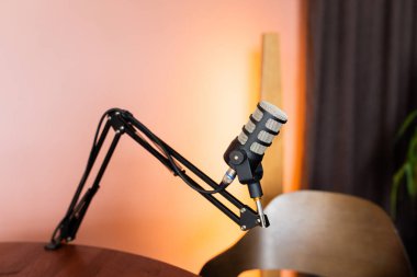 Pürüzsüz gümüş bir mikrofon, sıcak bir podcast stüdyosundaki ahşap bir masanın yanına yerleştirilmiş siyah ayarlanabilir bir standa monte edilir. Ortam davetkar ve profesyonelce..