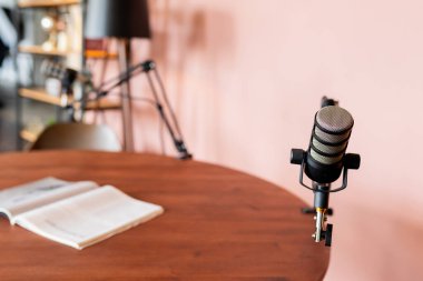 Bir podcast stüdyosunda ahşap bir masadaki açık bir defterin yanına yerleştirilmiş yüksek kaliteli bir mikrofon sergileniyor. Arkaplanda, sıcak bir atmosfere katkıda bulunan yumuşak pembe bir duvar yer alıyor..