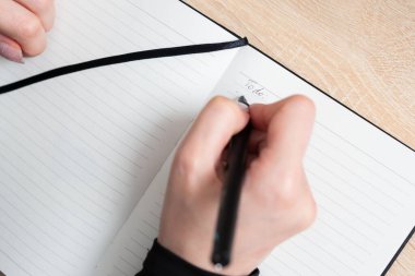 Bir kişi ahşap bir masada otururken siyah kalemle bir not defterine yapılacaklar listesi yazmaya odaklanır. Yumuşak doğal ışık çalışma alanını aydınlatıyor ve verimli bir atmosfer yaratıyor..