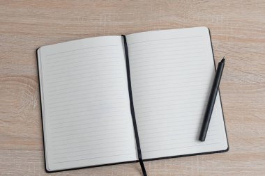 Boş sayfaları olan açık bir defter ahşap bir masada yatıyor. Yanında siyah bir kalem duruyor, yazarı fikir ve düşüncelerle doldurmaya davet ediyor..