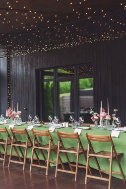 Büyüleyici bir düğün resepsiyonu yeşil masa örtüsüyle süslenmiş uzun bir masayı gösteriyor. Masada çiçekli süsler, cam eşyalar ve mum tutacakları var. Hepsi de parlak ışıkların altında..