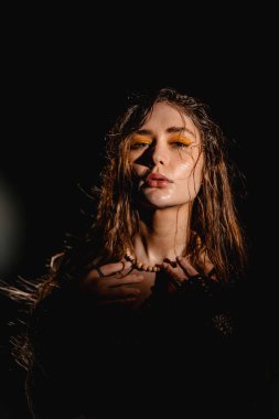 Bir kadın model, loş bir alanda, ıslak saçlar ve koyu sarı göz makyajıyla güvenle duruyor. Bir kolye tutuyor ve gizemli bir ortam yaratıyor..