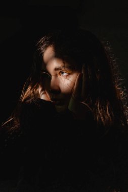 Genç bir birey düşünceli, yumuşak ışıkla aydınlanan ve yüzlerine karmaşık gölgeler düşen bakışlar atar. Karanlık arka plan, anın duygusal yoğunluğunu arttırır..