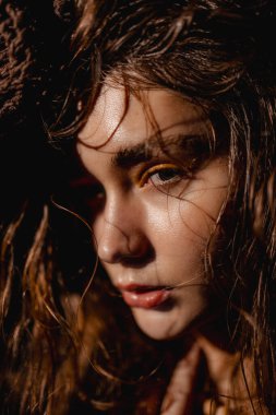 Islak saçı yüzüne doğru yavaşça düşerken genç bir kadın yoğun bir şekilde bakıyor. Altın göz makyajı, yumuşak ışıklandırmanın kapalı bir mekanda sıcak bir atmosfer yarattığı gibi onun özelliklerini vurgular..