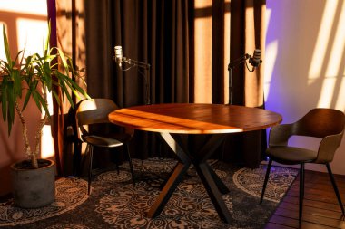 Sahne, bir yuvarlak ahşap masa, iki mikrofon ve röportaj için hazırlanmış sandalyelerin yer aldığı iyi ışıklandırılmış bir podcast alanını gösteriyor. Güneş ışığı perdelerin arasından süzülüp sıcak bir atmosfer yaratır..