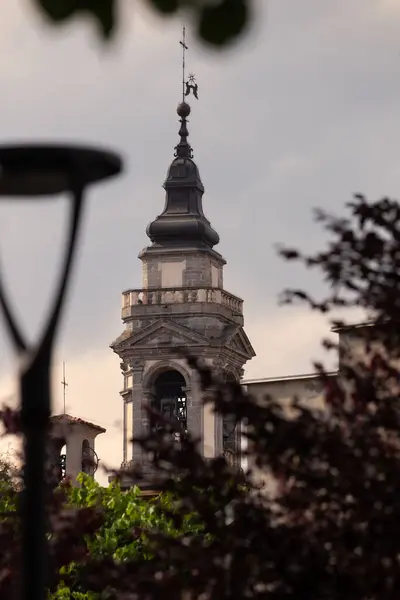 Yeşil yapraklarla çevrili ve kasvetli bir gökyüzüne kurulmuş tarihi bir çan kulesi. Detaylı mimari unsurlar çarpıcı bir tezat oluşturuyor, dingin ama dramatik görsel estetiği somutlaştırıyor..