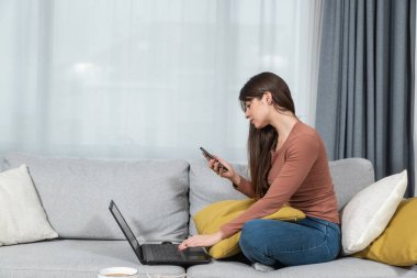 Kanepede oturan genç bir kadın akıllı telefonda promosyon kodlarını okuyor. İnternet sitesinde indirimli fiyata dizüstü bilgisayarda yazıyor..