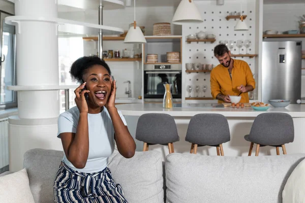 Young happy African American woman talking on smartphone feeling exited, winning vacation trip with her boyfriend who stands in the kitchen making a dinner. Lottery winning concept.