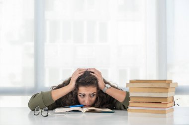 Kıvırcık saçlı genç okul ya da üniversite öğrencisi kızın başı ağrıyor ve saatlerce kitap okuduktan ve eğitim sınavına çalıştıktan sonra yorgun düşüyor. Endişe ve göz ağrısı konsepti.
