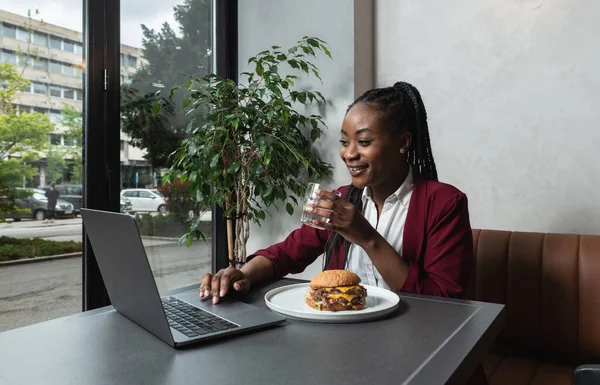 Young happy college exchange student or African American freelancer business woman having issue eating big fat tasty cheeseburger with melting cheese while she work on laptop computer in cafeteria on lunch break.