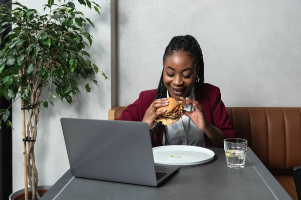 Young happy college exchange student or African American freelancer business woman having issue eating big fat tasty cheeseburger with melting cheese while she work on laptop computer in cafeteria on lunch break.