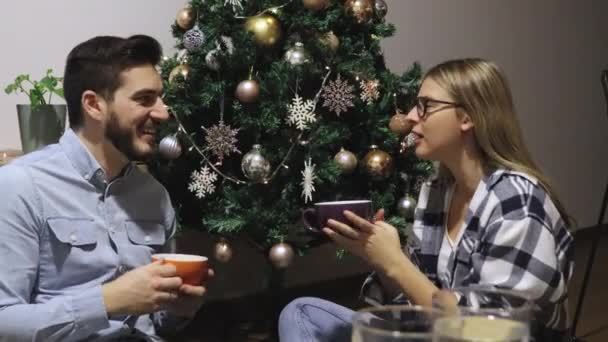 一对年轻夫妇坐在圣诞树前的地板上 装饰着新年装饰 一边喝咖啡或茶 一边享受着 一边聊天 男人和女人坐在一起开玩笑 在假日大笑 — 图库视频影像