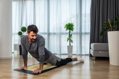 Kanserden kurtulan genç adam evde yoga antrenmanı yapıyor, kaslarını esnetiyor ve uzun süre acı ve hastalıkla mücadele ettikten sonra sağlıklı bir yaşam için egzersiz yapıyor. Meditasyon için ısınma egzersizleri.