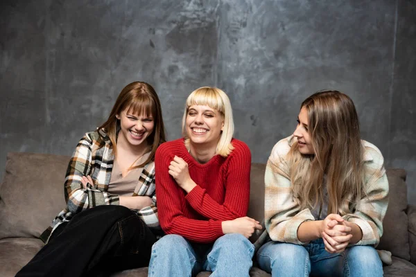 Three young female friends, roommates from college are sitting on the sofa and laughing because they are happy and successful in their lives.