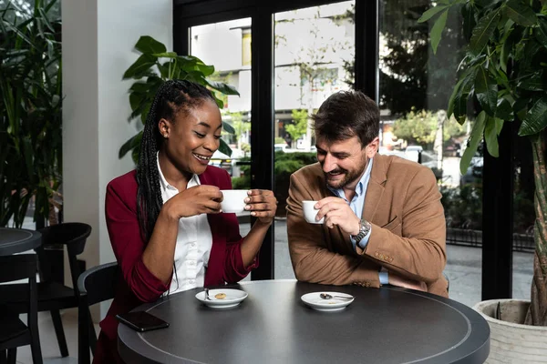 Young man and African American woman sitting in cafeteria drinking coffee flirting talking laughing and seducing outside working hours and colleague in restaurant away from busy street