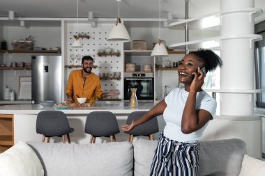 Young happy African American woman receive call she got the job she applied for at the company and her boyfriend smiling in the kitchen because now they will have more money for bills and better life