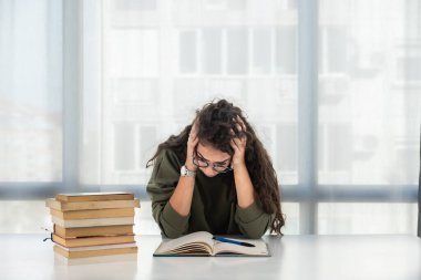 Young girl future law or economics student feeling nervous and anxious because she has a lot to study for her college entrance exam sitting with a pile of books on the table and has a headache