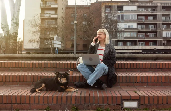 Young freelance businesswoman working outside on a laptop computer with takeaway coffee and her dog she adopted from a dog shelter. Web developer and graphic designer works out of the office